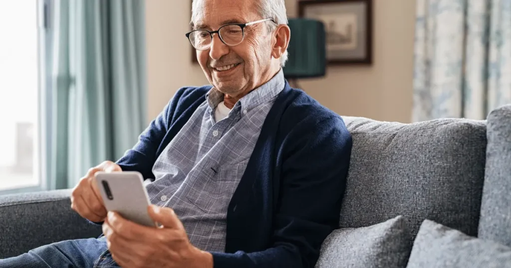 Äldre man med glasögon sitter bekvämt i en soffa och ler medan han använder sin smartphone.
