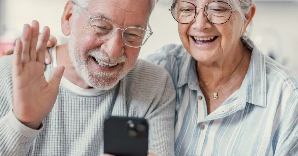 Äldre par med glasögon ler glatt medan de tittar tillsammans på en mobiltelefon och vinkar.