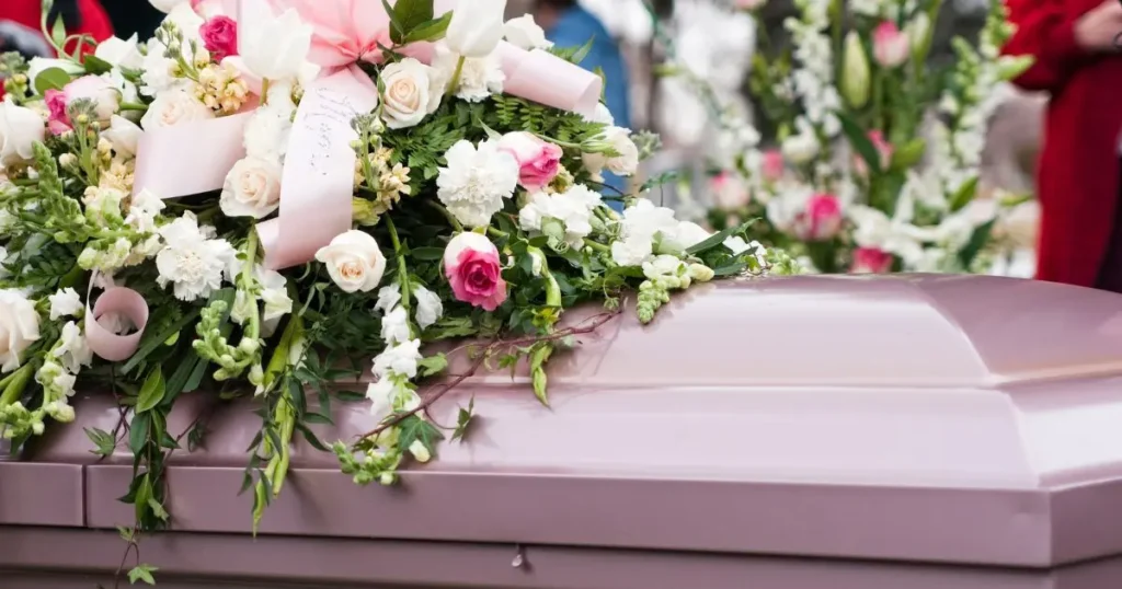 Vacker begravningskrans med vita och rosa rosor ligger på ett rosa kistlock under en ceremoni.