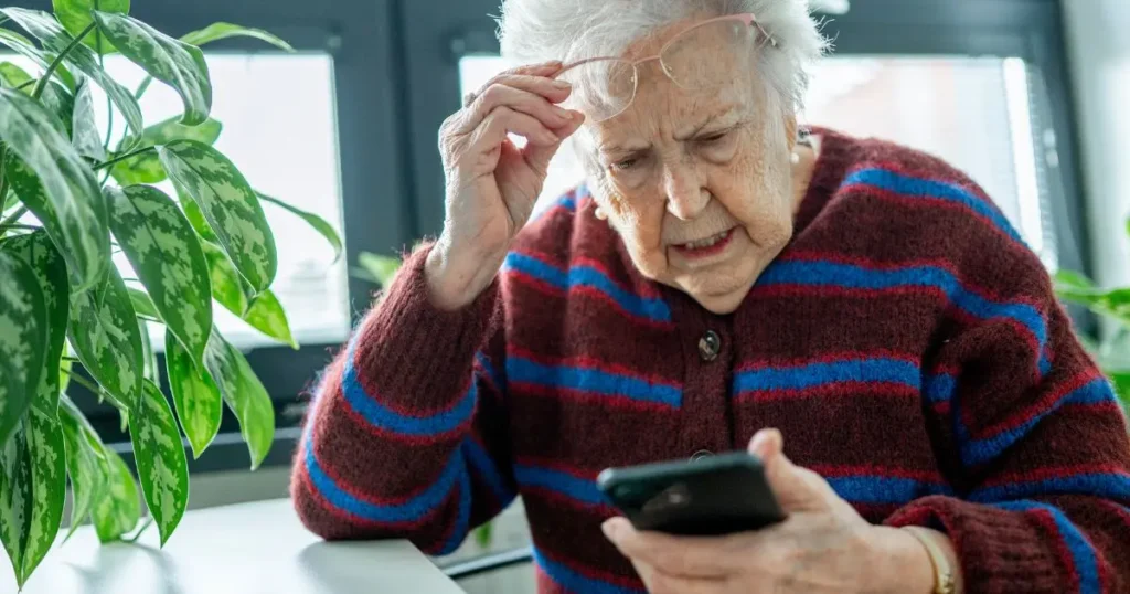 Äldre kvinna med glasögon tittar fundersamt och oroligt på sin mobiltelefon medan hon håller handen mot pannan.