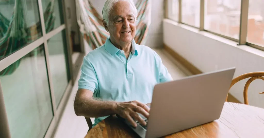 Äldre man med vitt hår sitter leende vid ett träbord och använder en laptop i ett ljust rum med stora fönster.