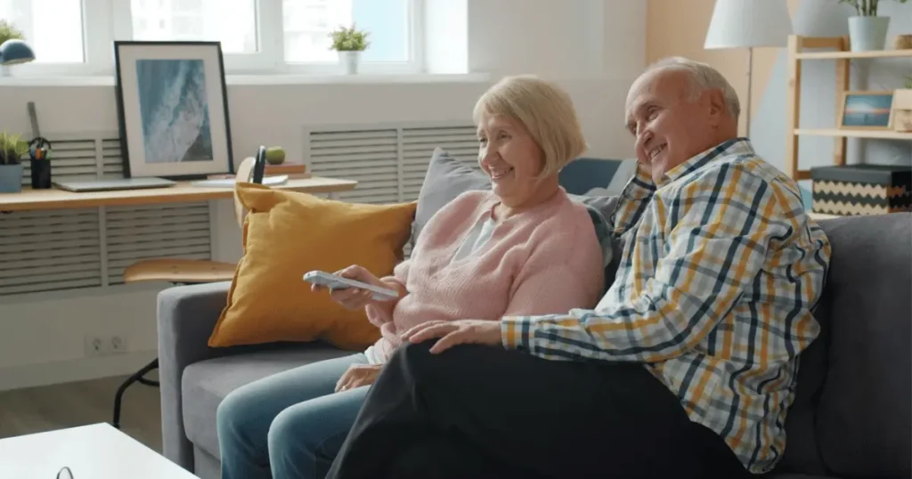 Äldre par sitter nöjda i en soffa. Kvinnan håller en fjärrkontroll medan de tittar mot TV:n.