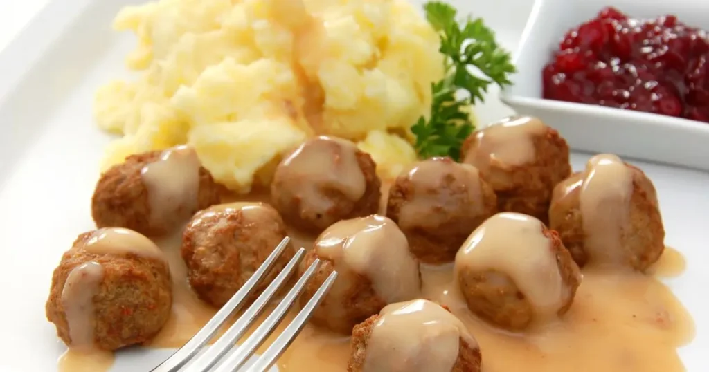 Tallrik med köttbullar i gräddsås, potatismos och rårörda lingon med en gaffel i förgrunden.