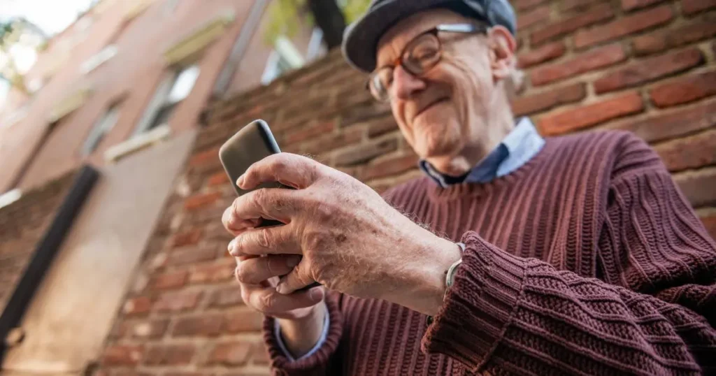 Äldre man med glasögon och keps ler medan han håller och använder en mobiltelefon.
