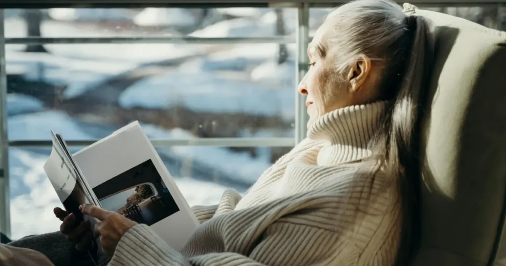 Äldre kvinna med långt vitt hår sitter bekvämt i en fåtölj och läser en tidning vid ett stort fönster.