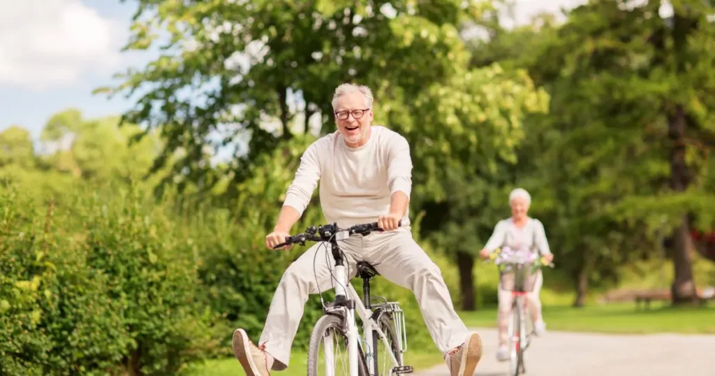 Glädjande äldre man med glasögon cyklar leende på en stig i en grönskande park.